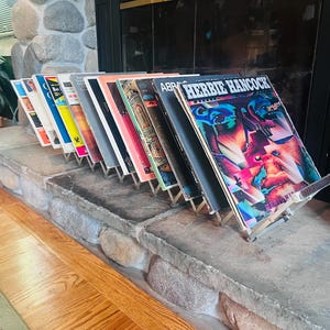 May include: A collection of vinyl record albums displayed in a metal stand. The front album cover features the text Herbie Hancock and displays a vibrant, abstract graphic design with shades of blue, purple, and pink against a dark background.