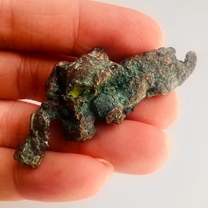 May include: A rough, irregularly shaped piece of copper ore with a dark brown and green patina. The mineral specimen has a textured surface and is held in a hand against a white background.