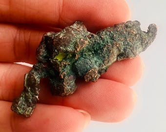 May include: A rough, irregularly shaped piece of copper ore with a dark brown and green patina. The mineral specimen has a textured surface and is held in a hand against a white background.