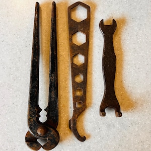 May include: Three vintage metal tools with a rusty brown finish. The tools include a pair of pliers, a multi-size wrench, and a small open-ended wrench. The tools are arranged on a light-colored surface.