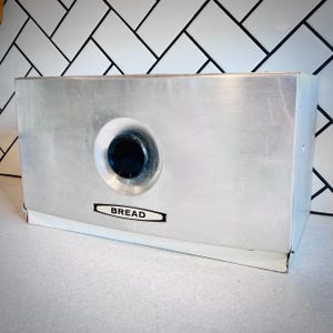 May include: A rectangular brushed metal bread box with a smooth finish. The front panel features a circular black knob centered in a recessed area and a horizontal black and white label with the word BREAD printed in clear capital letters.
