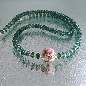 Pu&ograve; includere: Una collana di pietre preziose verdi con un ciondolo floreale bianco e rosa. La collana &egrave; composta da piccole perle rotonde e ha una chiusura argentata.