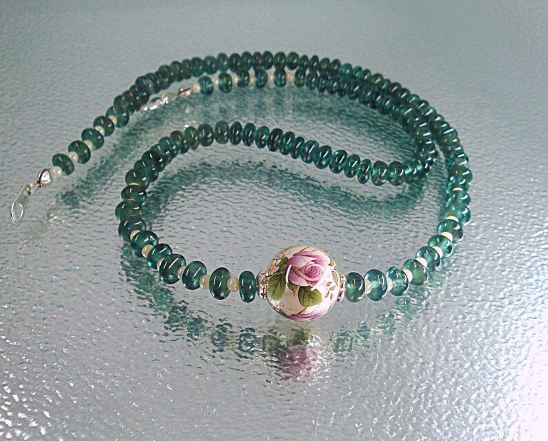 Pu&ograve; includere: Una collana di pietre preziose verdi con un ciondolo floreale bianco e rosa. La collana &egrave; composta da piccole perle rotonde e ha una chiusura argentata.