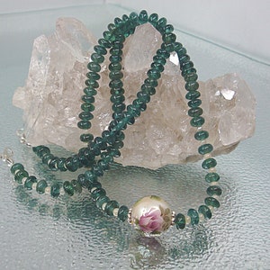 Pu&ograve; includere: Una collana di perline di pietre preziose verdi con una perla di porcellana bianca con un design di rosa rosa.