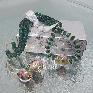 Pu&ograve; includere: Una collana di perline di vetro verde con orecchini con motivo floreale in una scatola regalo argentata con un nastro bianco.