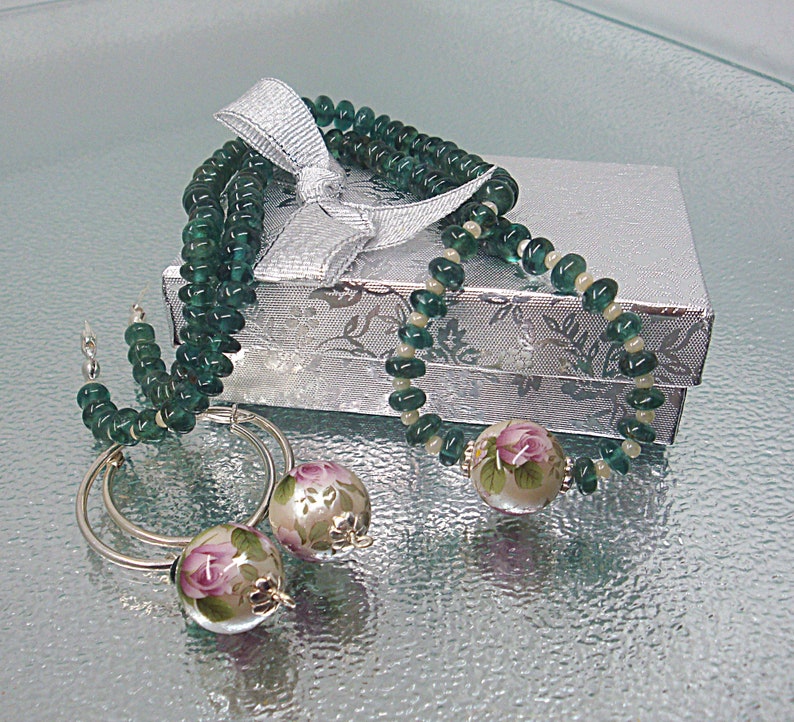 Pu&ograve; includere: Una collana di perline di vetro verde con orecchini con motivo floreale in una scatola regalo argentata con un nastro bianco.