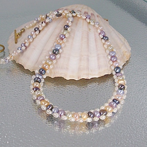 Può includere: Una collana di perle a più fili con chiusura dorata. La collana presenta una varietà di colori di perle, tra cui bianco, lavanda, grigio e pesca. La collana è esposta su una grande conchiglia.