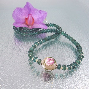 Pu&ograve; includere: Una collana di pietre preziose verdi con un unico grande ciondolo rotondo con un motivo floreale. La collana &egrave; appoggiata su una superficie argentata con un fiore di orchidea rosa.