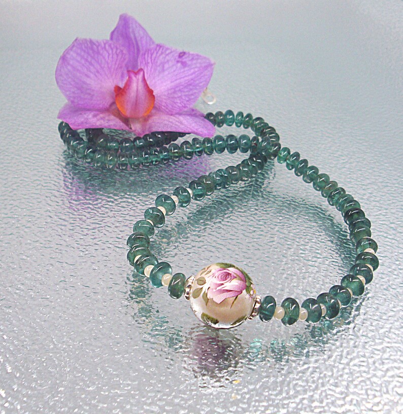 Pu&ograve; includere: Una collana di pietre preziose verdi con un unico grande ciondolo rotondo con un motivo floreale. La collana &egrave; appoggiata su una superficie argentata con un fiore di orchidea rosa.