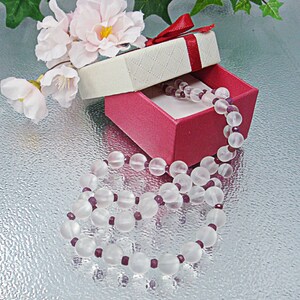 Peut inclure: Un arrangement de fleurs blanches et roses se trouve à côté d'une boîte cadeau rose avec un couvercle blanc. La boîte est ouverte et un collier de perles blanches et violettes se trouve à l'intérieur.