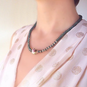Pu&ograve; includere: Una collana di perle di porcellana verde e bianca con un motivo floreale. La collana &egrave; indossata da una persona con una blusa rosa a pois.