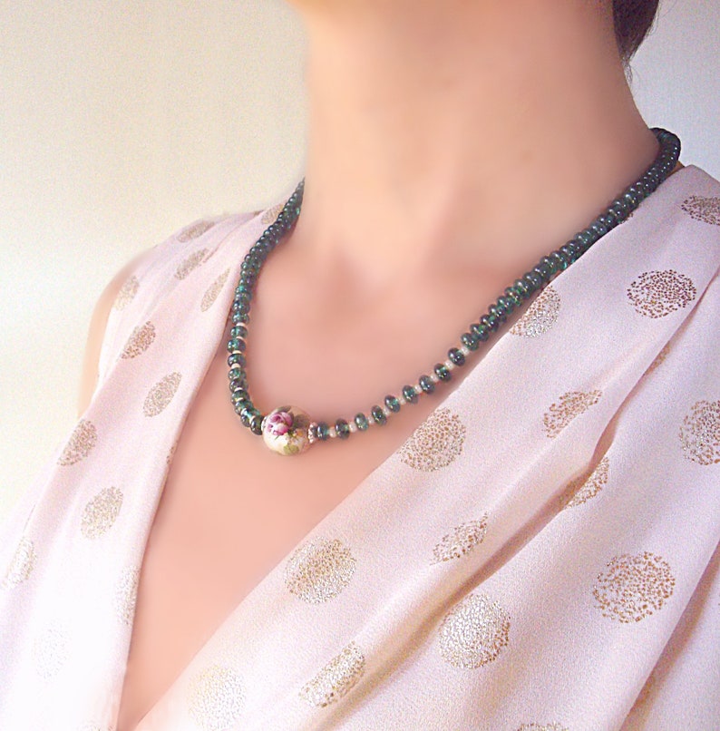 Pu&ograve; includere: Una collana di perle di porcellana verde e bianca con un motivo floreale. La collana &egrave; indossata da una persona con una blusa rosa a pois.