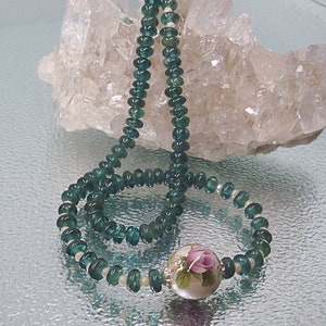 Pu&ograve; includere: Una collana di pietre preziose verdi con un ciondolo floreale bianco e rosa. La collana &egrave; esposta su un letto di cristalli di quarzo bianchi.