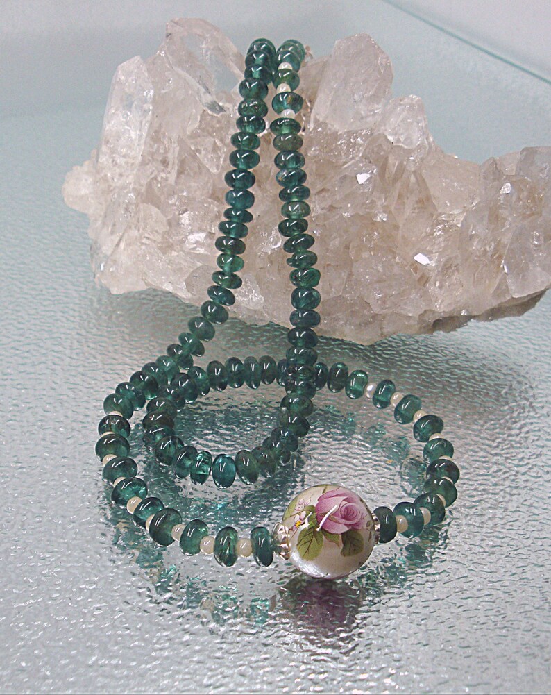 Pu&ograve; includere: Una collana di pietre preziose verdi con un ciondolo floreale bianco e rosa. La collana &egrave; esposta su un letto di cristalli di quarzo bianchi.