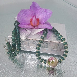Pu&ograve; includere: Una collana di perline verdi e bianche con un ciondolo floreale. La collana &egrave; esposta su una scatola regalo argentata con un'orchidea rosa in cima.