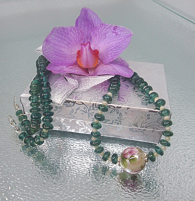 Pu&ograve; includere: Una collana di perline verdi e bianche con un ciondolo floreale. La collana &egrave; esposta su una scatola regalo argentata con un'orchidea rosa in cima.
