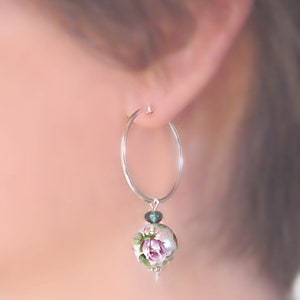 Pu&ograve; includere: Orecchini a cerchio d'argento con un ciondolo floreale. Il ciondolo presenta una rosa rosa con foglie verdi su uno sfondo bianco. Il ciondolo &egrave; accentato da una piccola pietra preziosa verde.