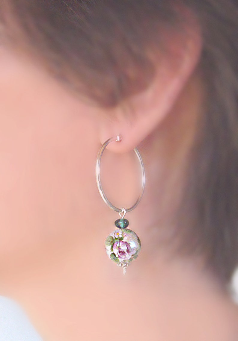 Pu&ograve; includere: Orecchini a cerchio d'argento con un ciondolo floreale. Il ciondolo presenta una rosa rosa con foglie verdi su uno sfondo bianco. Il ciondolo &egrave; accentato da una piccola pietra preziosa verde.