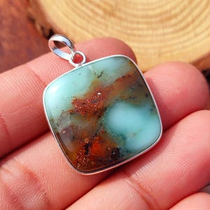 May include: Square pendant with a silver-coloured bail. The pendant features a light blue and brown stone with a silver-coloured border. The stone has a unique pattern of colours and textures.