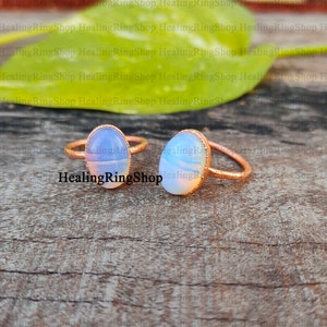 May include: Two rings with oval, opalescent gemstones and copper-toned bands. The stones display a mix of white and blue colours. The rings are presented on a wooden surface. The text "HealingRingShop" is visible.