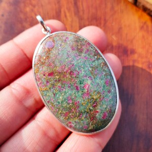 Puede incluir: Un colgante de forma ovalada con una piedra pulida de textura moteada en tonos verde oscuro, magenta vibrante y motas doradas metálicas. La piedra está engastada en un marco de metal plateado liso y sencillo con una pequeña anilla.