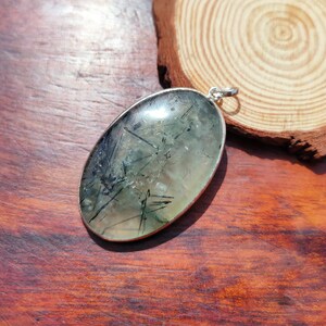 May include: A silver pendant featuring a clear oval-shaped stone with black inclusions. The stone is set in a silver bezel and has a polished finish.