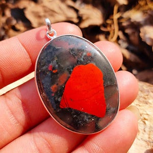 May include: A silver pendant featuring a large, oval-shaped red jasper stone with black and grey inclusions. The stone is set in a silver bezel and has a polished finish.