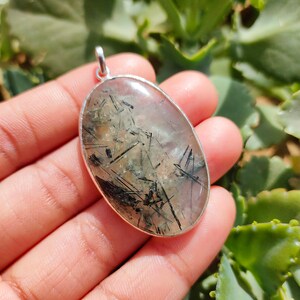 May include: Silver pendant featuring a clear quartz stone with black tourmaline inclusions. The stone is oval shaped and has a smooth, polished finish.
