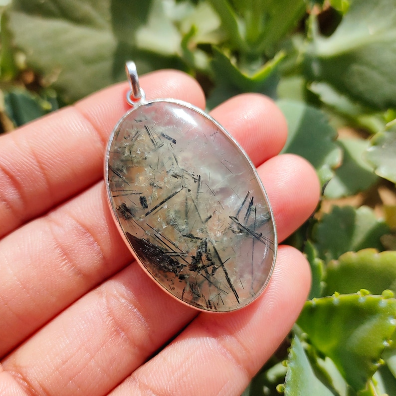 May include: Silver pendant featuring a clear quartz stone with black tourmaline inclusions. The stone is oval shaped and has a smooth, polished finish.
