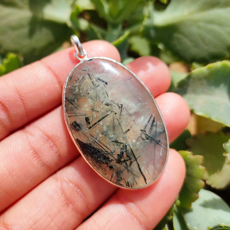 May include: Silver pendant featuring a clear quartz stone with black inclusions. The stone is oval shaped and has a smooth, polished finish.
