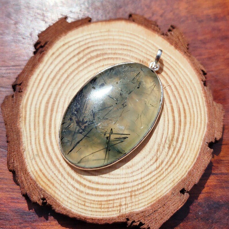 May include: A silver pendant featuring a green and black oval gemstone with a silver bail. The gemstone has a translucent appearance with black inclusions.