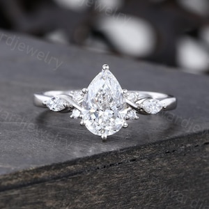 May include: A silver-toned ring featuring a central pear-cut clear stone, flanked by two smaller marquise-cut stones on each side, with additional tiny round stones set along the band, creating a multi-stone arrangement on a polished metal band.