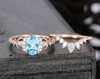 May include: A rose gold engagement ring set with a large oval-shaped blue topaz gemstone surrounded by smaller diamonds. The matching wedding band features a row of pear-shaped diamonds.