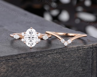May include: Rose gold engagement ring set featuring an oval-cut center stone and accent diamonds. The set includes a matching V-shaped band with small diamonds. The rings are displayed on a dark wooden surface.