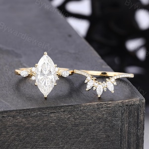 Può includere: Un set di anelli in oro composto da un anello di fidanzamento con una grande pietra centrale taglio marquise affiancata da pietre più piccole, abbinato a una fede nuziale curva decorata con pietre taglio marquise e rotonde.