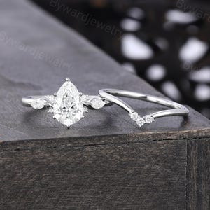 May include: A silver-toned engagement ring set featuring a pear-cut center stone flanked by two marquise-cut side stones, accompanied by a matching V-shaped wedding band adorned with three small round stones at the center point of the band.