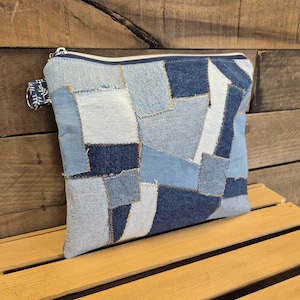 May include: A rectangular zippered pouch constructed from various shades of blue denim fabric arranged in a patchwork pattern. The pouch features a silver-toned metal zipper closure and a small fabric loop attached to the side with a white leaf print.