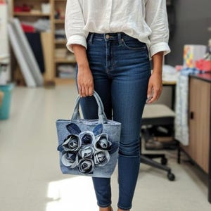 May include: A denim tote bag with fabric rose embellishments. The bag is a light blue denim with darker blue roses. The tote has two handles and is being held by a person wearing a white shirt and blue jeans.
