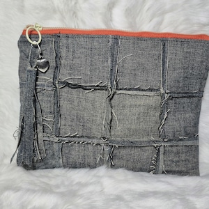 May include: A denim patchwork clutch with an orange zipper and a silver heart charm. The gray denim bag has a rectangular shape with visible stitching and frayed edges, giving it a rustic, handmade appearance.