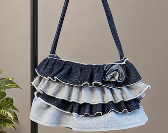 May include: A denim shoulder bag featuring three horizontal tiers of ruffled fabric. The ruffles alternate between dark indigo and light blue denim washes. A circular denim rosette accent is attached to the top right corner of the bag.