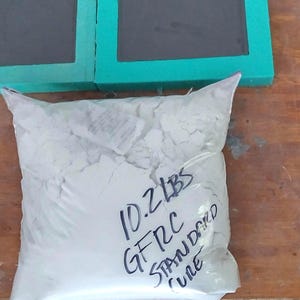 以下が含まれることがあります： 10.2 LBS GFRC STANDARD CUREと書かれた白い粉末入りの透明なビニール袋と、中央がダークグレーで青、緑、ベージュの枠が付いた3つの正方形の型が、木製の表面に並べられています。