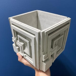 May include: A square, light gray concrete planter featuring a raised, geometric maze-like pattern on its exterior sides. The vessel has a hollow, open top and a textured, matte finish, showcasing a structured, architectural design with sharp, clean lines.