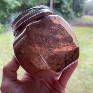 Puede incluir: Un frasco de plástico transparente de forma cuadrada lleno de un polvo fino de color marrón rojizo oscuro. El recipiente cuenta con una tapa metálica de rosca con un borde estriado. El polvo en su interior se ve denso y compacto.