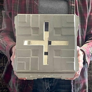 May include: A light grey, square-shaped concrete lantern featuring a geometric, recessed relief pattern on its front face and a central cross-shaped cutout that allows light to pass through the hollow interior of the structure.