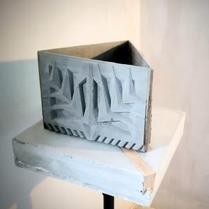 Puede incluir: Una maceta triangular de hormigón con un exterior gris que presenta un patrón geométrico en relieve de líneas nítidas y angulares, junto con una serie de muescas verticales a lo largo del borde inferior, creando una superficie texturizada.