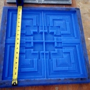 May include: A square blue silicone mold featuring a geometric, stepped, maze-like pattern. A yellow measuring tape is placed vertically across the mold, indicating a length of approximately 17 inches from the top edge to the bottom edge.