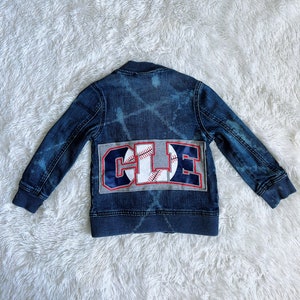 May include: A dark blue denim jacket with a faded design. The jacket features a patch with the letters "CLE" in red, white, and blue, with a baseball graphic. The jacket is designed for children.