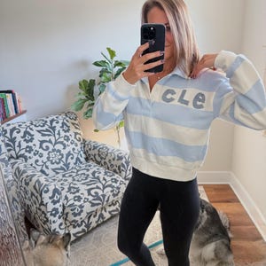 May include: A light blue and white striped sweatshirt with the letters "CLE" on the front. The sweatshirt has a collared neckline. The person is wearing black leggings and brown sandals. A dog is visible in the mirror.
