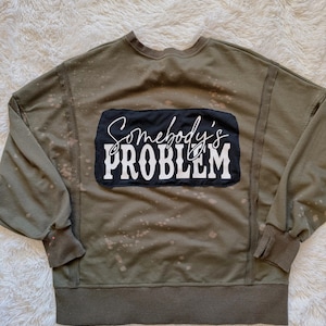 May include: An olive green sweatshirt with the text "Somebody's PROBLEM" in white script and block letters on a black rectangle. The sweatshirt has a crew neck, long sleeves, and a distressed, bleached appearance.