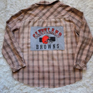 May include: A long-sleeved, button-down shirt with a plaid pattern in shades of brown and pink. A gray patch on the back features the words "Cleveland Browns" with a football helmet logo in red and black. The shirt is made of a soft, comfortable material.